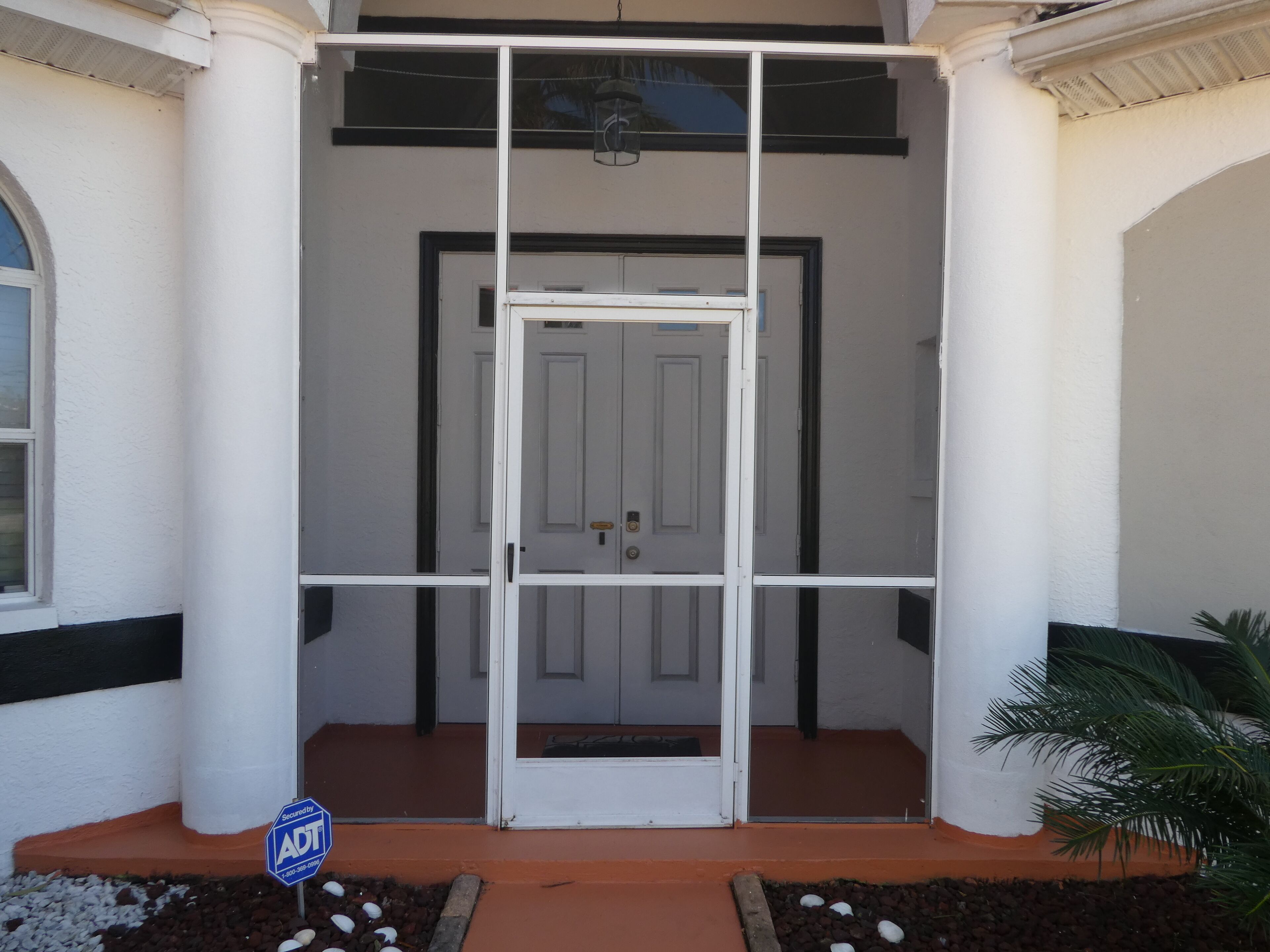 A white building with a central door, flanked by two columns, and a small garden area with pebbles and a palm tree.