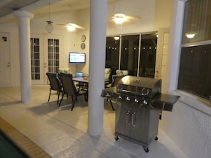 A covered outdoor area with a grill, dining table, chairs, and a television screen.