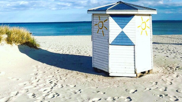 A beach hut with a simple design, featuring a sun symbol on its side, located on a sandy beach with footprints leading up to it. - Feel-good apartment StrandGut für Ostseegenießer, Hs. Atlantic (Grömitz)