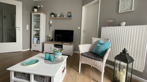 A modern living room with a white entertainment unit, a wicker chair with a cushion, a small table with decorative items, and a TV. - Feel-good apartment StrandGut für Ostseegenießer, Hs. Atlantic (Grömitz)