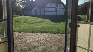 A view through a glass door of a house with a thatched roof, a paved lawn, and a brick pathway leading to the entrance. - Comfortable holiday house for 7 persons with sauna, 110 sqm, in the south of Rügen (Zudar/Rügen)
