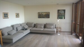 Een woonkamer met een grijze, hoekbank, twee ingelijste foto’s aan de muur en een vloerlamp.
