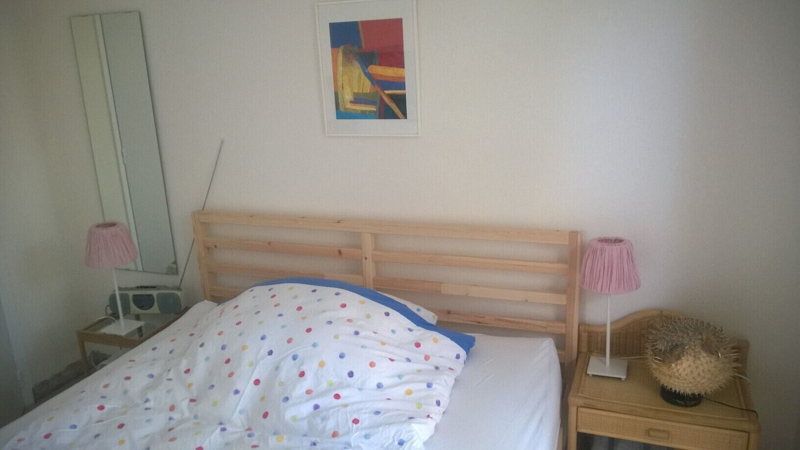 A bedroom with a wooden bed frame, a white bedspread with colorful polka dots, a bedside lamp, a radio, and a decorative item on the nightstand.