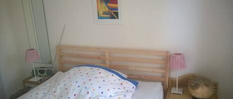 A bedroom with a wooden bed frame, a white bedspread with colorful polka dots, a bedside lamp, a radio, and a decorative item on the nightstand.