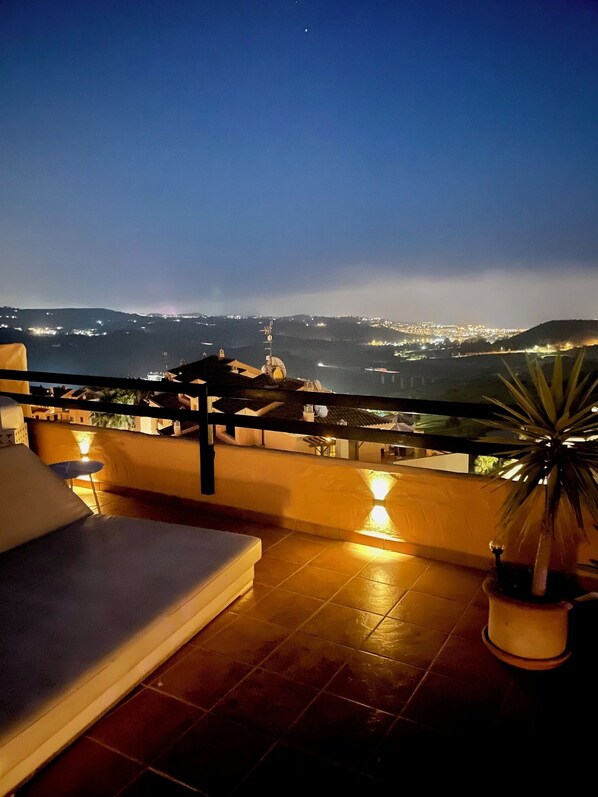 A terrace with a view of a city at night, including a pool area and a potted plant.
