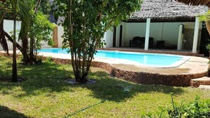 A pool area with a thatched roof structure, surrounded by greenery and a stone wall. - Villa LeoMar: Exclusive vacation home with pool & garden, 350 m from the beach (Diani Beach)