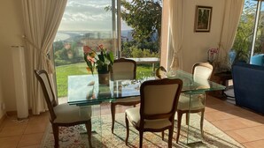Comedor con mesa de vidrio, cuatro sillas y vistas a un jardín y paisaje distante a través de amplios ventanales.