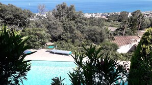 Una piscina con dos tumbonas, rodeada de exuberante vegetación y vista al mar y edificios a lo lejos.