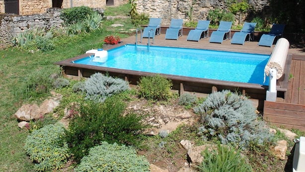 Une piscine entourée de chaises longues, elle-même bordée d’un jardin où poussent différentes espèces de plantes, avec une maison en pierre en arrière-plan.