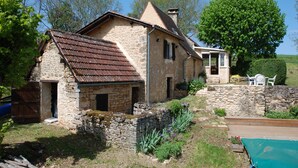 Une maison en pierre avec un toit en pente, une terrasse en bois et un jardin agrémenté de plantes et d’un arbre.