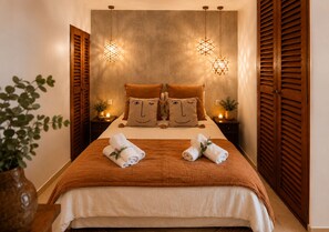 A neatly made bed with a brown blanket, two pillows with a face design, and two towels with a flower. There are two hanging lamps with a geometric pattern, a wooden door with louvered panels, and a plant on the side. - Exceptional cottage with private garden, 100m from Tahiti beach &  St Tropez 2mn (Ramatuelle)