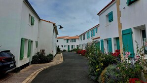 Uma rua estreita ladeada por edifícios brancos com persianas verdes, um carro estacionado e diversas flores.