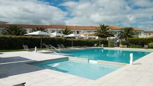 Piscina com espreguiçadeiras e guarda-sóis, cercada por um sebedo e palmeiras, com edifícios ao fundo.