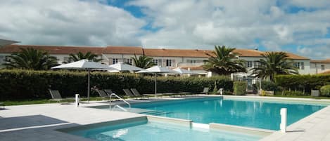 A swimming pool with lounge chairs and umbrellas, surrounded by a hedge and palm trees, with buildings in the background.