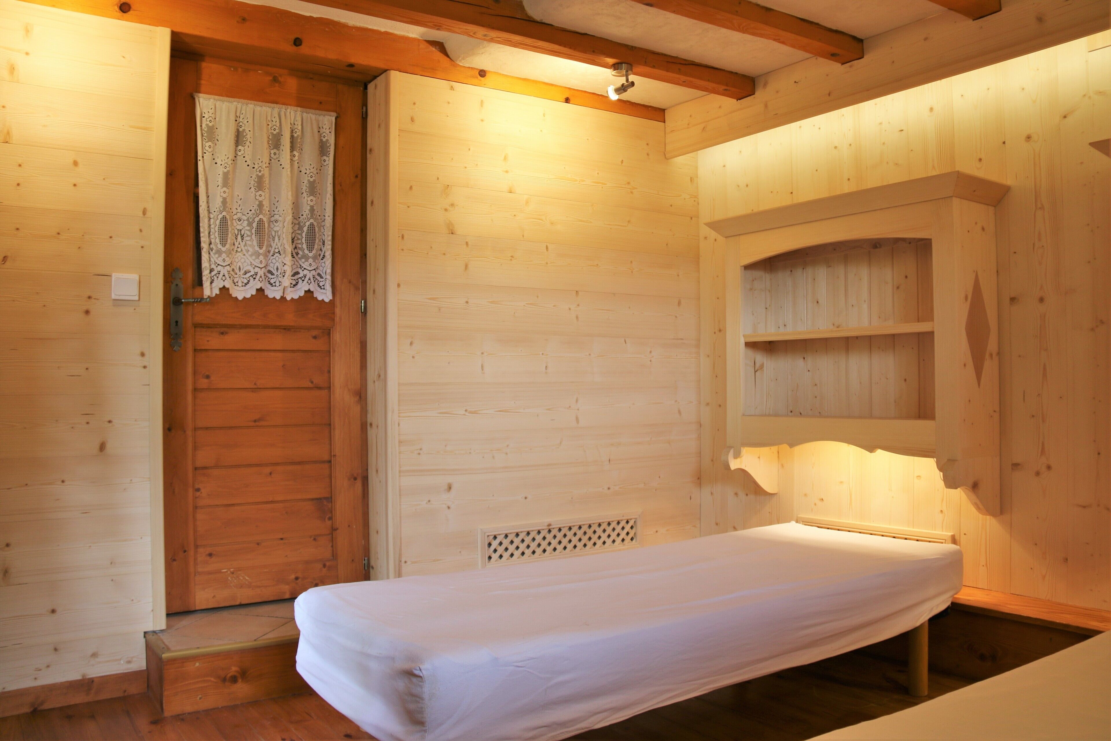 A wooden cabin room with a single bed, a wooden door with a lace curtain, and a shelf unit.