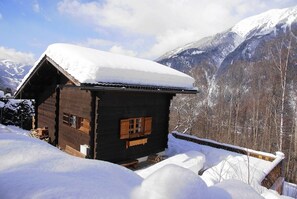 En stuga av trä med ett snötäckt tak, omgiven av ett snöigt landskap och berg.