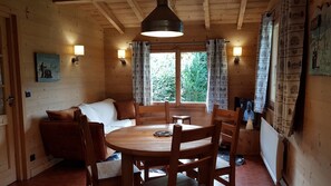 A cozy cabin interior with a wooden dining table, chairs, a sofa, and a large window with curtains. - Two cozy chalets with mountain views near Annecy and Aravis (Thônes)