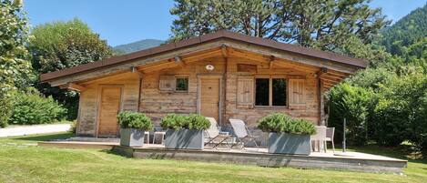 Eine Holzhütte mit geneigtem Dach, großen Fenstern und einer Veranda mit Sitzgelegenheit.