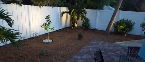 Un patio trasero con cerca blanca, un árbol pequeño y un área de patio con mesa y sillas.