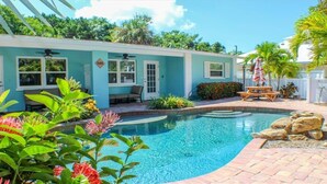 A pool area with a blue building, outdoor seating, and tropical plants.