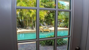 A view through a window of a swimming pool surrounded by palm trees and lounge chairs.