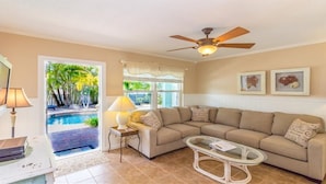 A living room with a beige sofa, a glass coffee table, a ceiling fan, and a view of a pool through a glass door.