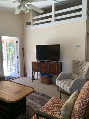 Een woonkamer met een televisie op een houten kast, een fauteuil met kussens, een houten salontafel en een kamerkleed met een patroon.