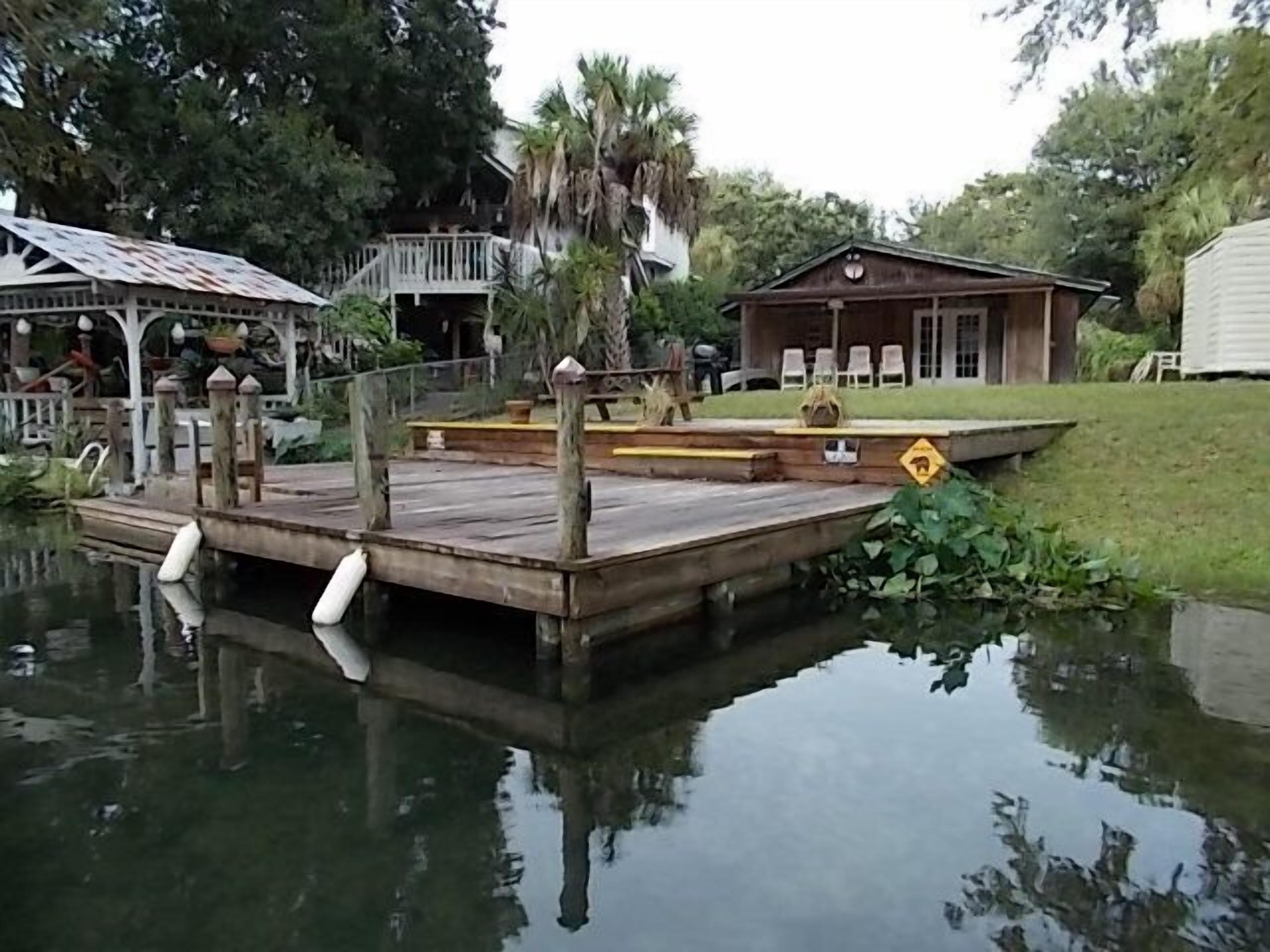 Un muelle de madera se extiende sobre aguas tranquilas, rodeado de vegetación exuberante y un pequeño edificio con porche.