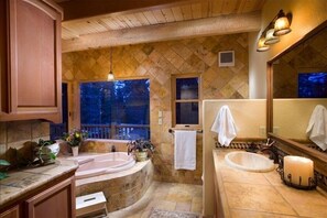 A bathroom with a large bathtub, a sink, and a mirror. There are towels hanging on the wall and a window with a view of trees.
