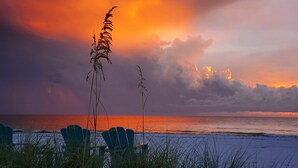 A beach scene with chairs and grass in the foreground, and a sunset over the ocean. - Dunes, Daisies, Surf, Sand, Sunsets...Historic BUTLERHOUSE,  Pass-a-Grille (St. Pete  Beach)