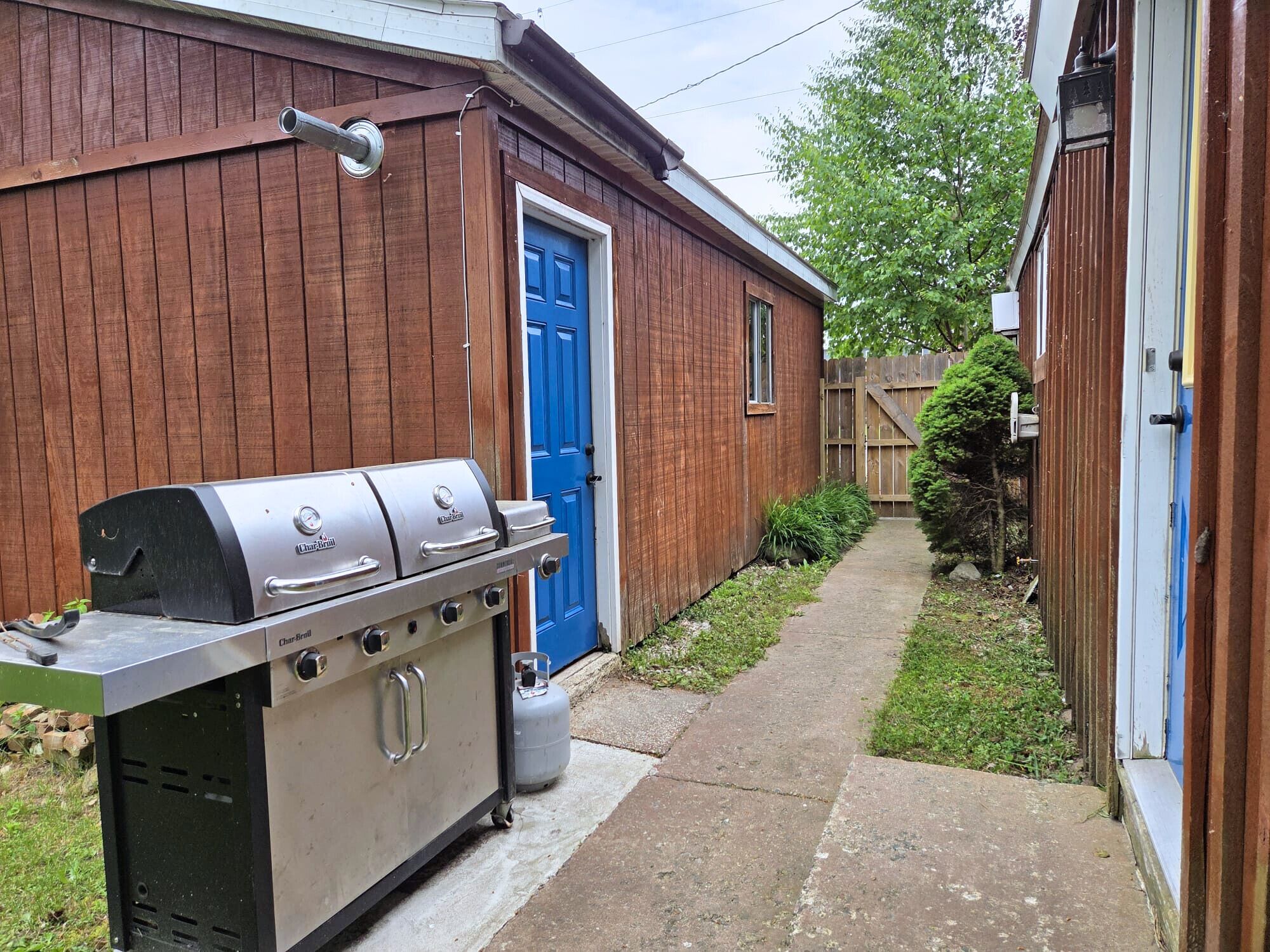 A barbecue grill outside a wooden building with a blue door, a concrete pathway, and a small garden area.
