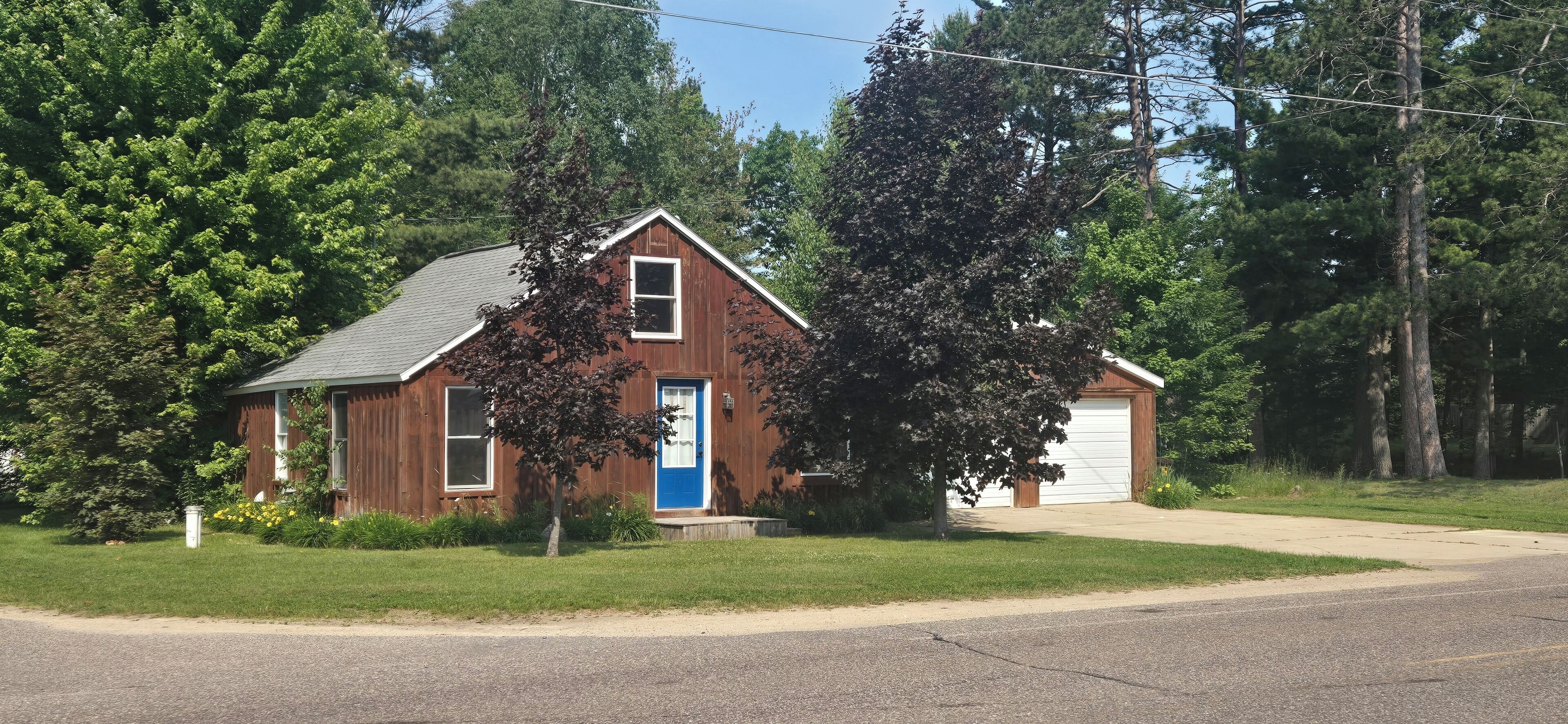 A two-story wooden house with a blue garage door, surrounded by trees and a grassy yard.