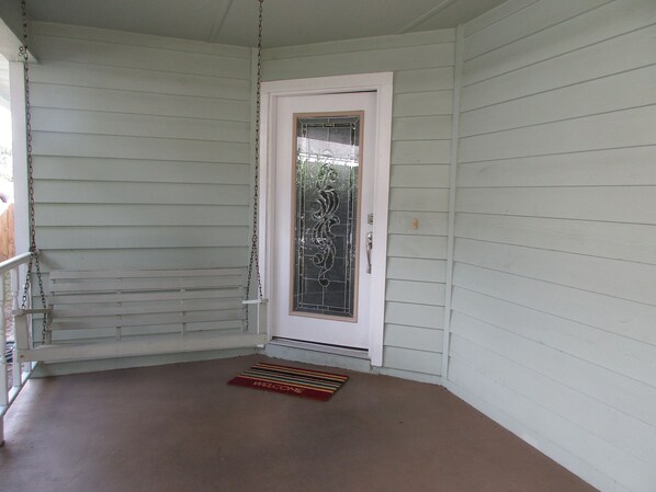 Eine weiße Tür mit Fensterglas und Metallbeschlägen, ein kleiner Podestschritt führt zur Tür, und eine Fußmatte liegt auf der Veranda.
