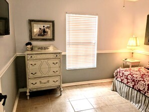 Une chambre à coucher avec une commode, un lit recouvert d’une couvre-lit à motifs floraux, une fenêtre avec des stores et une lampe sur une table de chevet.