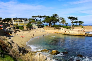 A coastal beach scene with clear waters, rocky shoreline, and a building with a balcony overlooking the beach.