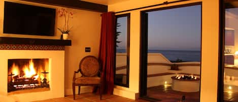 A cozy living room with a fireplace, a comfortable chair, and a view of the ocean through large sliding doors.