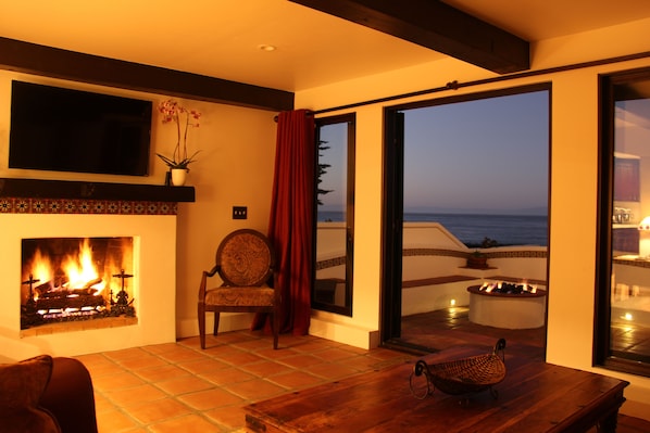 A cozy living room with a fireplace, a comfortable chair, and a view of the ocean through large sliding doors.
