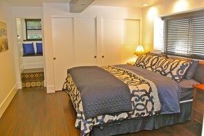 A bedroom with a large bed, a nightstand, a lamp, a window with blinds, a door, and a small staircase with a decorative box.