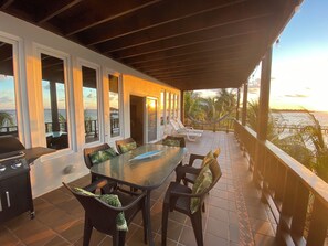A covered outdoor dining area with a glass table and chairs, overlooking a beach and ocean at sunset.