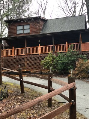 Cozy cabin with a balcony and porch, surrounded by lush trees and a paved path.