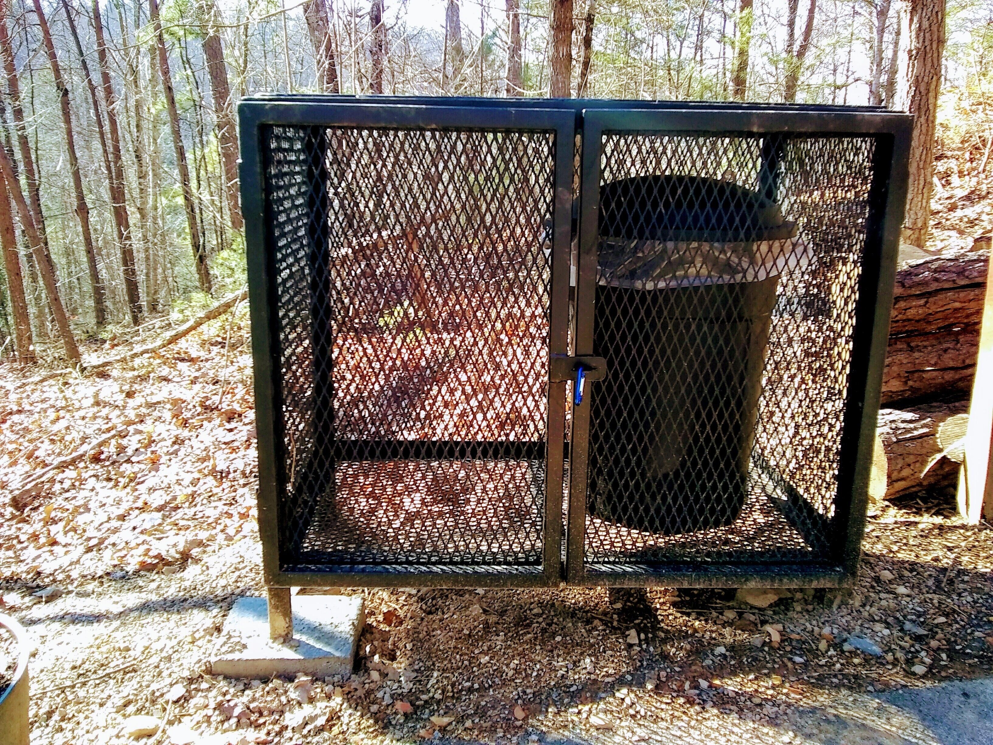 A metal outdoor trash bin with a mesh cover, placed on a gravel surface.