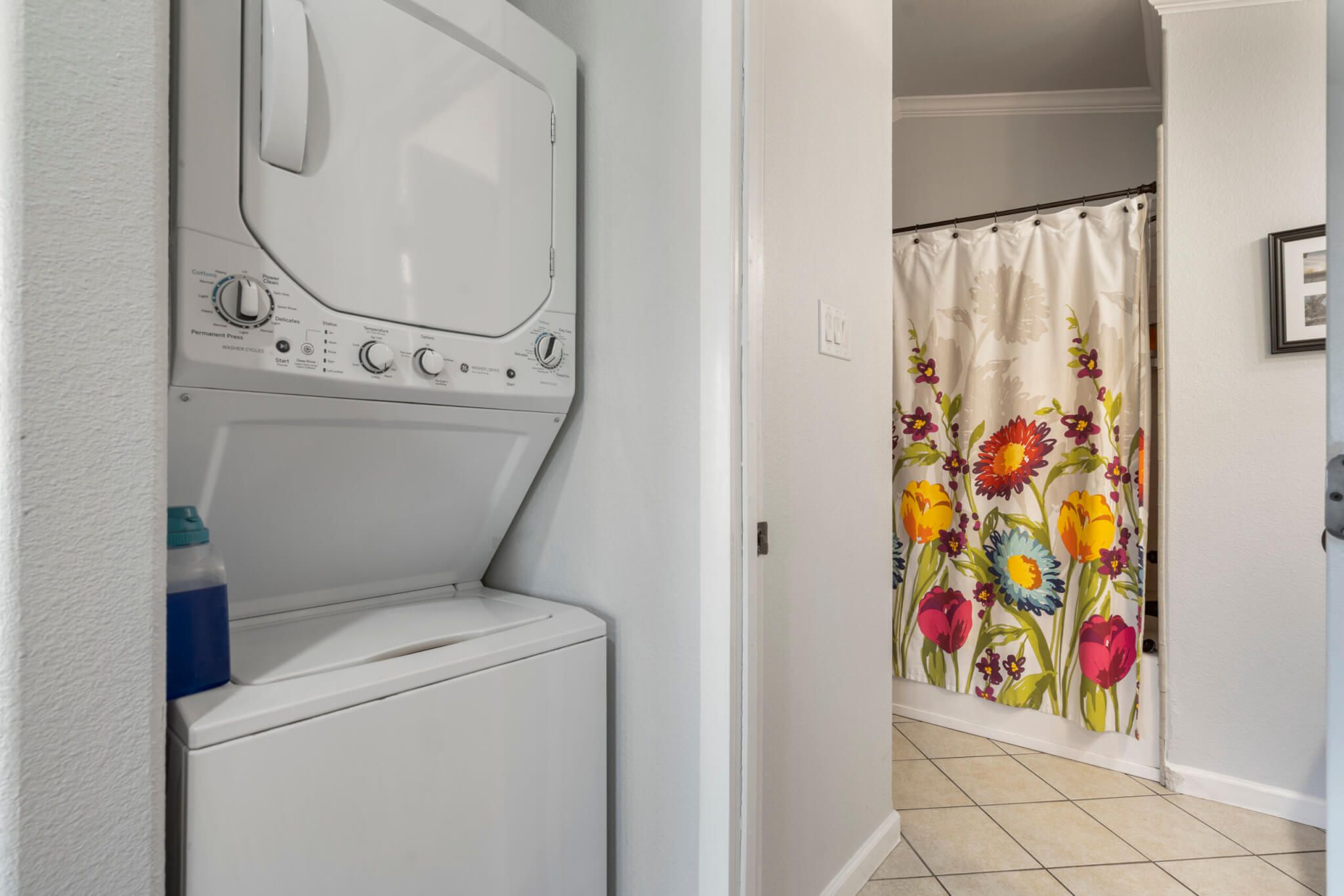 Un área de lavandería con lavadora y secadora de carga frontal, cortina de ducha con estampado floral y piso de baldosas.