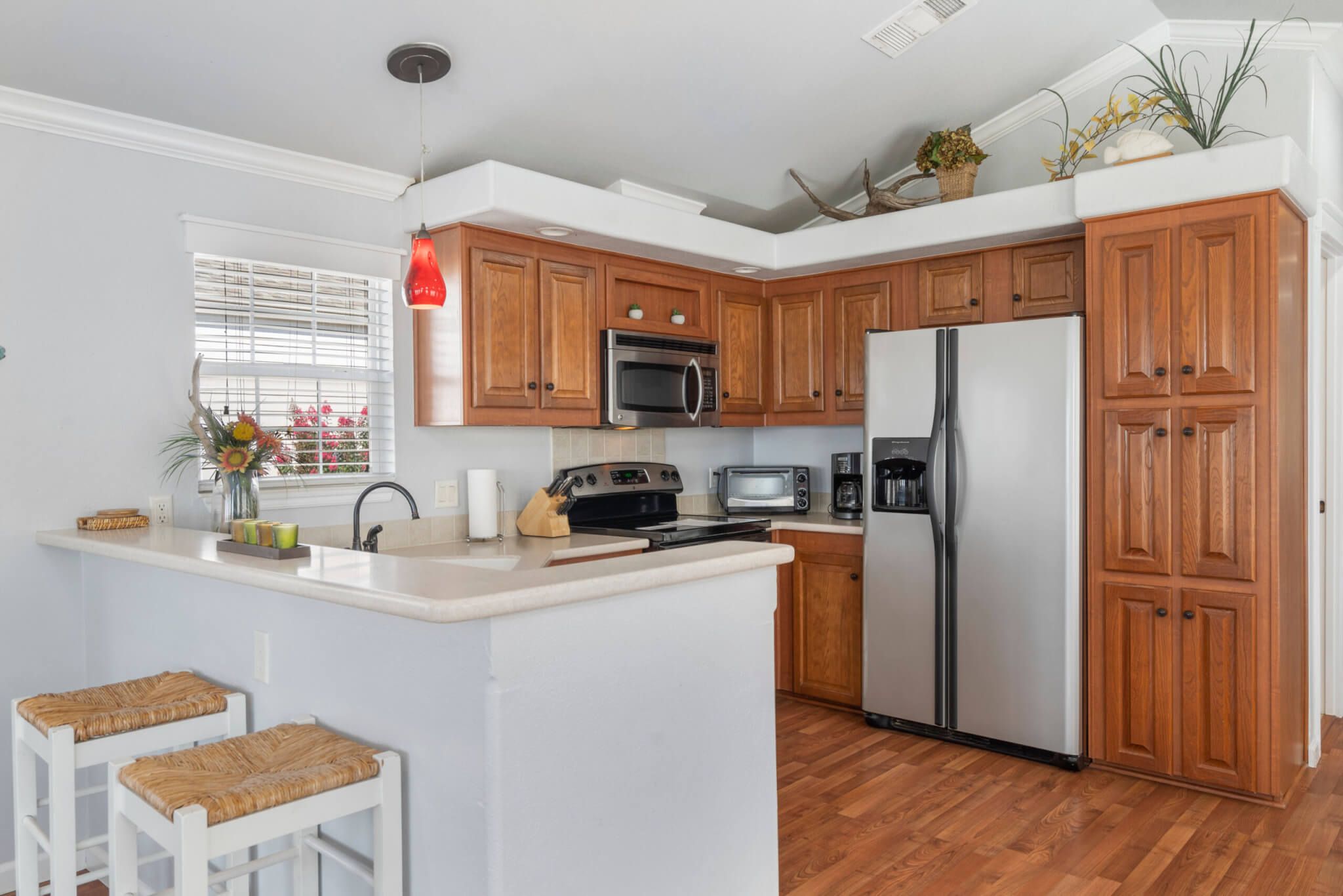 A kitchen with wooden cabinets, a stainless steel refrigerator, a microwave, and a toaster. There are two white stools and a window with blinds.