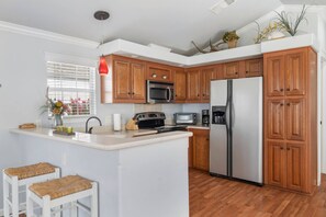 A kitchen with wooden cabinets, a stainless steel refrigerator, a microwave, and a toaster. There are two white stools and a window with blinds. - Rockport at CrossTimbers Marina (Sperry)