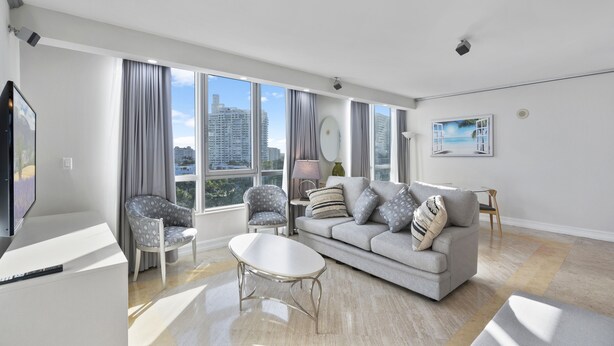 Ein modernes Wohnzimmer mit einem grauen Sofa, zwei Sesseln, einem Couchtisch und einem Fernseher. Große Fenster bieten Blick auf die Stadt.