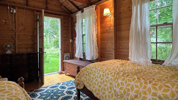 A wooden cabin bedroom with a bed, a dresser, a fan, and a window with curtains. - Peaceful hideaway on Westport Point (Westport)