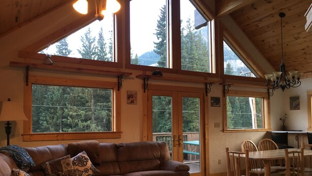 Un salón amplio con un sofá de cuero marrón, una mesa de comedor de madera y sillas, y ventanas grandes que ofrecen vistas a una zona boscosa.