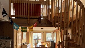 A spacious loft-style room with a wooden staircase, a dining area, and a stone fireplace.