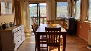 A dining area with a wooden table and chairs, a kitchen counter with tea sets, and large windows with curtains.