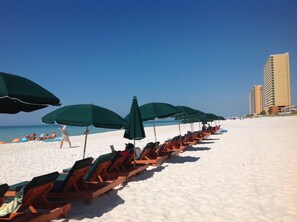 A beach with rows of wooden lounge chairs and green umbrellas, a clear blue sky, and a tall building in the background. - Unique, Elegant And Beautiful 17th Floor Condo At Emerald Isle (Panama City Beach)